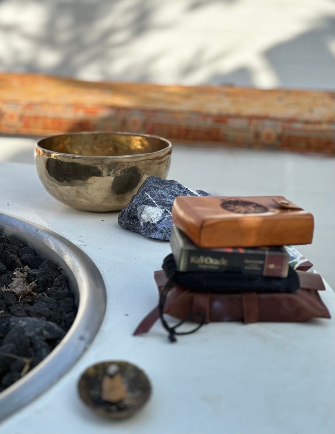 Sunlit outdoor patio meditation scene: brass singing bowl and raw dark crystal on a white table beside a lava-rock fire pit, with stacked leather pouches and a small wooden box.