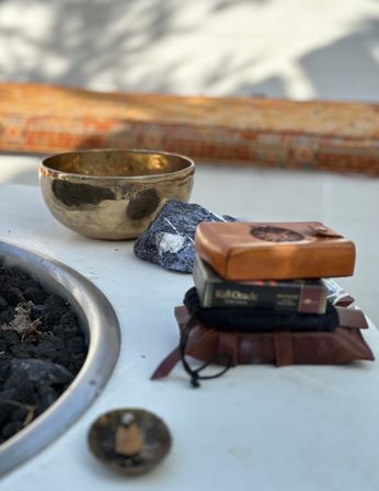 Sunlit outdoor patio meditation scene: brass singing bowl and raw dark crystal on a white table beside a lava-rock fire pit, with stacked leather pouches and a small wooden box.