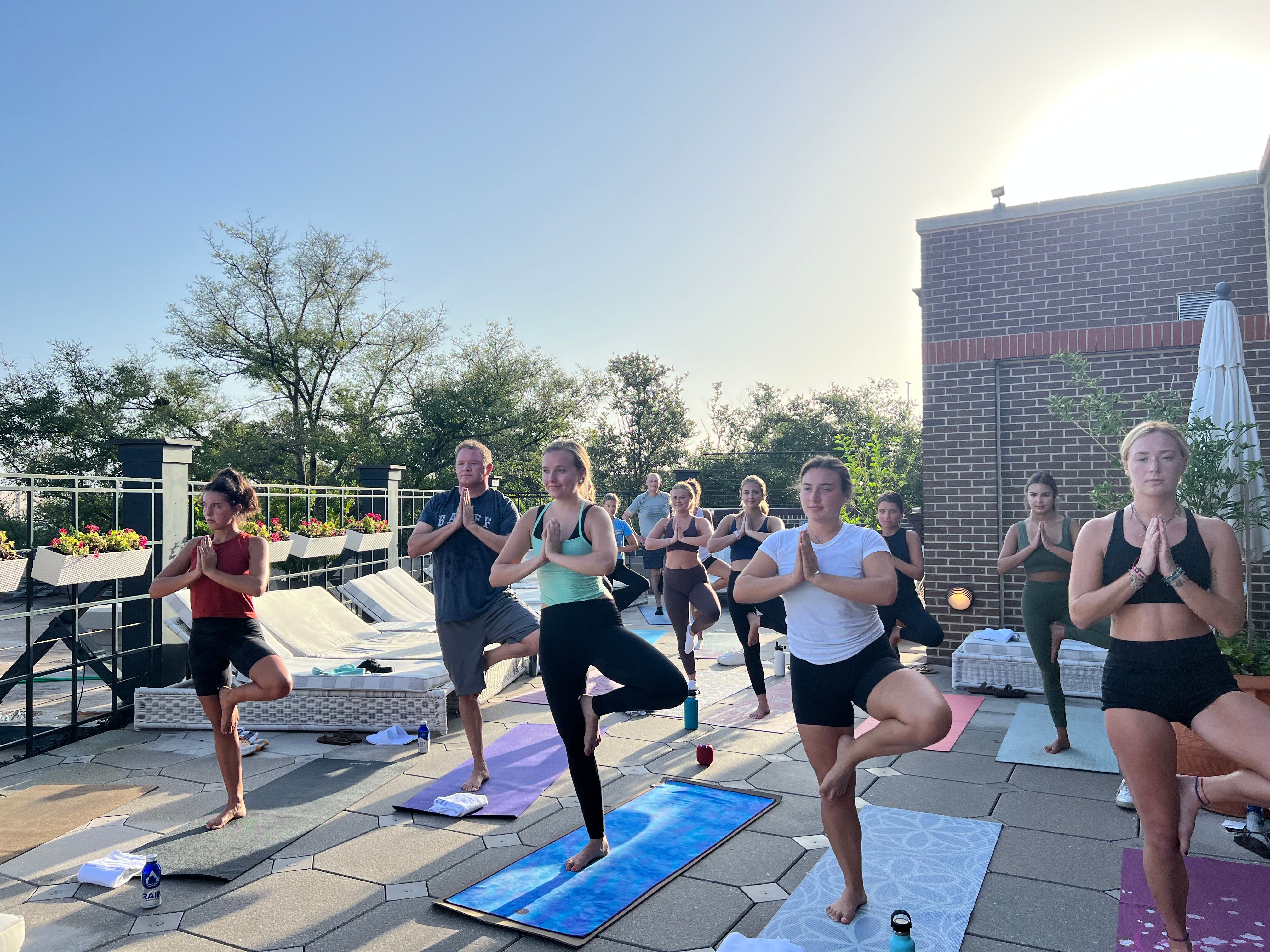 Sunlit rooftop yoga class at sunrise on a terrace, participants balancing in tree pose on colorful mats near lounge chairs and planters.