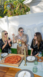 Sunny backyard bachelorette party — bride-to-be in satin robe pops sparkling wine while friends cheer, pizza and place settings on wooden table with white fence and palm trees in background.