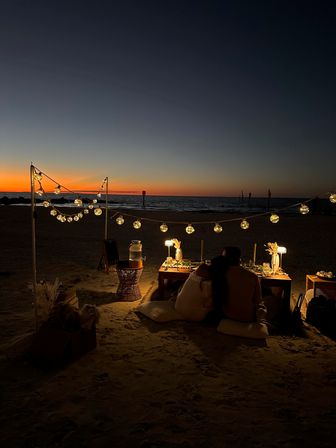 Cozy romantic beach picnic at sunset on the oceanfront, string globe lights over a low dining table set for two with cushions on the sand.