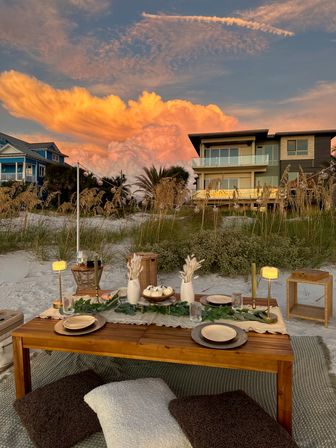 Coastal sunset beach picnic with a low wooden table, cushions, candles and tableware on white sand dunes, beachfront homes and dramatic orange clouds
