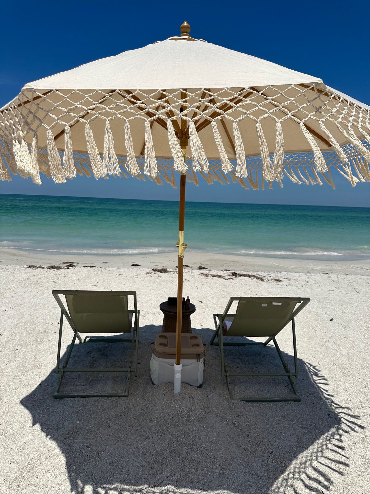 White tasseled beach umbrella shading two lounge chairs on powdery white sand with turquoise ocean and clear blue sky beyond.