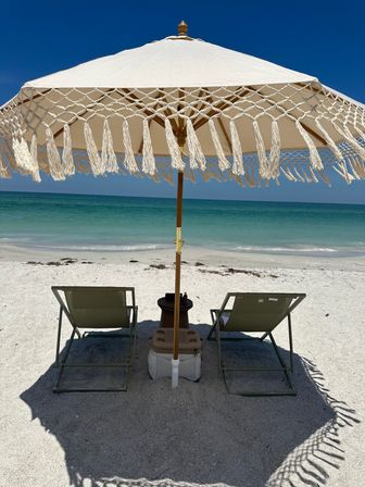 White tasseled beach umbrella shading two lounge chairs on powdery white sand with turquoise ocean and clear blue sky beyond.