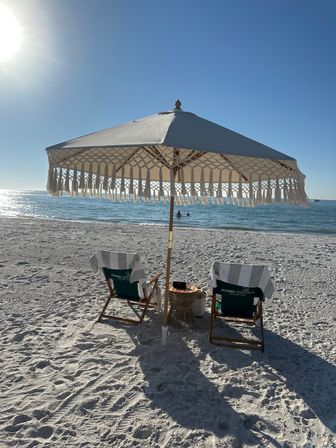 Sunny beach scene with a fringed umbrella shading two striped lounge chairs and a small side table on white sand, calm blue ocean and swimmers on the horizon.
