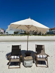 Two wooden beach chairs with striped towels under a fringed white umbrella beside a cooler on a sandy beach, with coastal houses and a deep blue sky in the background.