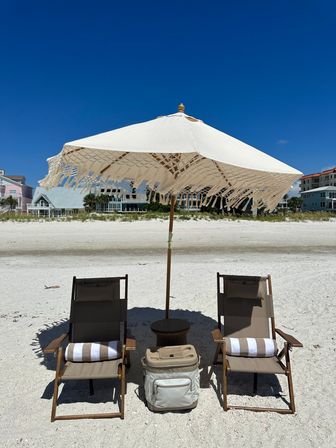 Two wooden beach chairs with striped towels under a fringed white umbrella beside a cooler on a sandy beach, with coastal houses and a deep blue sky in the background.
