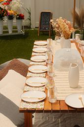 Sunny backyard garden picnic setup with a low wooden table lined with white plates and gold cutlery on floral napkins, iridescent glasses, dried-flower centerpieces in vases, cozy floor cushions and a chalkboard sign by buckets of blooms.