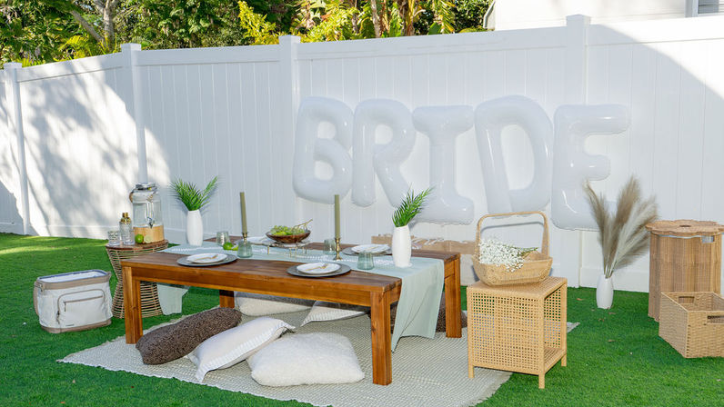 Outdoor backyard bridal picnic on green lawn with low wooden table, cozy floor cushions, rattan baskets, white 'BRIDE' balloons and white fence backdrop