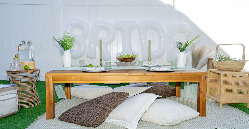 Backyard bridal picnic: low wooden table with place settings, white "BRIDE" balloons, floor cushions, greenery vases and wicker decor on grass