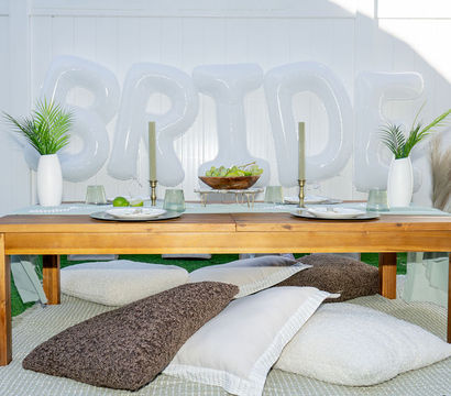 Backyard bridal picnic: low wooden table with place settings, white "BRIDE" balloons, floor cushions, greenery vases and wicker decor on grass