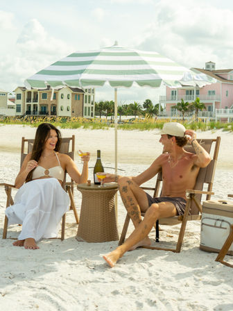 Couple toasting with drinks under a green-striped beach umbrella on a white-sand beach in front of pastel coastal homes