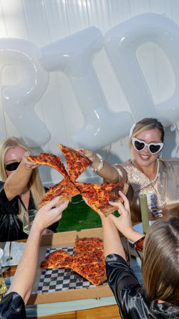 Group of friends clinking large pepperoni pizza slices over an open pizza box at an outdoor backyard bachelorette party with white 'BRIDE' balloons and sunglasses