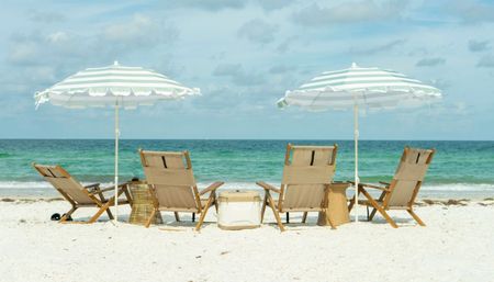 Four wooden lounge chairs and two green-and-white striped beach umbrellas on white sand facing turquoise ocean under a partly cloudy sky, relaxed seaside scene.
