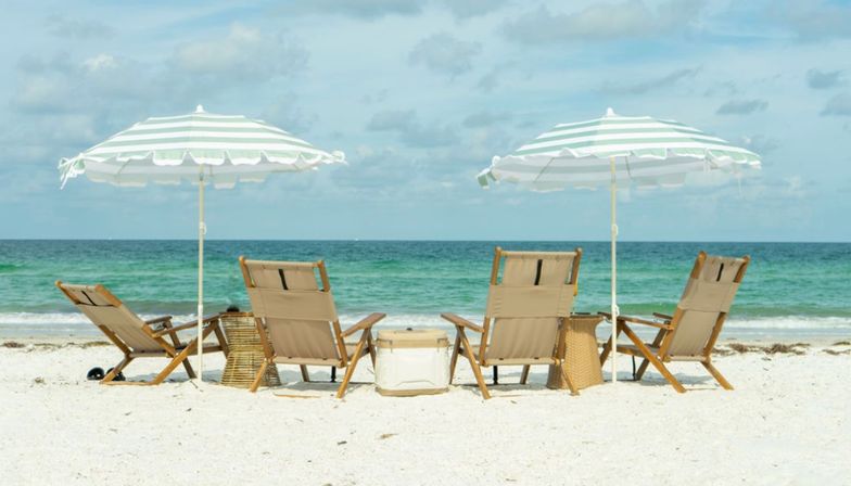 Four wooden lounge chairs and two green-and-white striped beach umbrellas on white sand facing turquoise ocean under a partly cloudy sky, relaxed seaside scene.