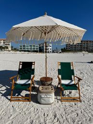 Boho fringe beach umbrella shading two green wooden lounge chairs with cooler and wicker table on a white-sand beach in front of beachfront condos under a clear blue sky
