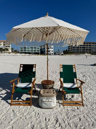 Boho fringe beach umbrella shading two green wooden lounge chairs with cooler and wicker table on a white-sand beach in front of beachfront condos under a clear blue sky