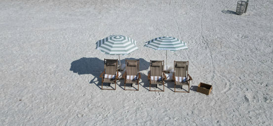Aerial view of four wooden lounge chairs under green-and-white striped umbrellas on a pristine white-sand beach, empty and sun-ready with a nearby wicker basket and distant trash bin.