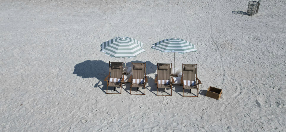 Aerial view of four wooden lounge chairs under green-and-white striped umbrellas on a pristine white-sand beach, empty and sun-ready with a nearby wicker basket and distant trash bin.