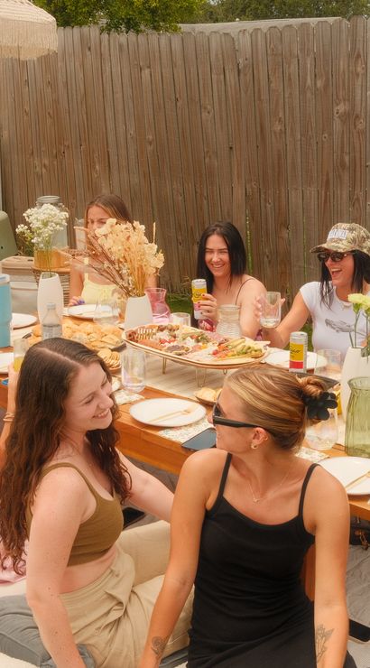 Sunny backyard brunch picnic — friends seated on cushions around a low wooden table on grass with a charcuterie board, pastries, drinks and floral vases under an umbrella by a wooden fence.