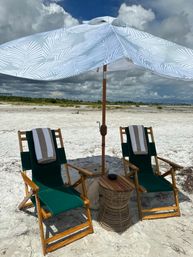 Two wooden green canvas beach chairs with striped towels under a patterned umbrella on a sandy beach beside a wicker side table, dramatic cloudy sky overhead
