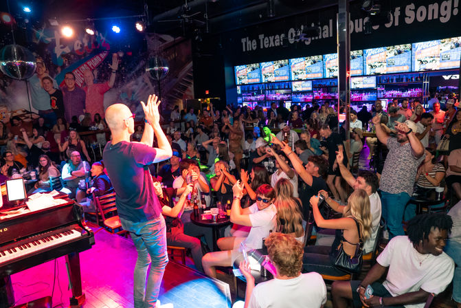 Crowded Texas piano‑bar nightlife scene with a performer clapping on stage, cheering patrons at tables, colorful stage lights, disco balls and a busy backlit bar.