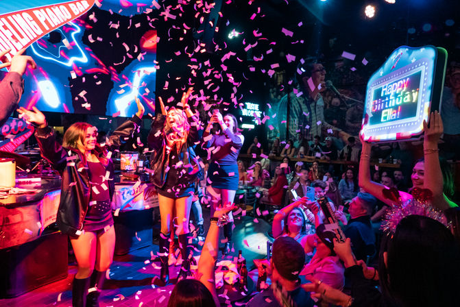 Neon-lit piano bar birthday party with confetti raining down on dancing performers while a cheering crowd captures the celebration on phones