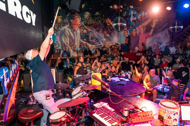 Energetic piano bar scene from the stage: performer raising a drumstick while a lively crowd cheers under disco balls, with keyboards, drums, microphones and a colorful mural backdrop.