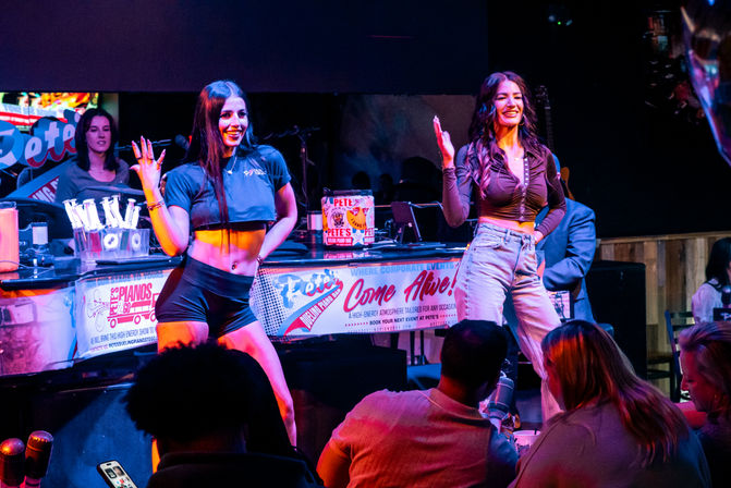 Two performers smiling and waving onstage at a lively dueling piano bar with colorful stage lights, band gear, and a seated audience enjoying live music nightlife.