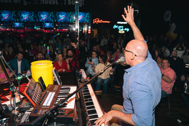 Energetic nightlife scene: musician at a keyboard sings with hand raised to a cheering crowd in a neon-lit, packed live music bar.