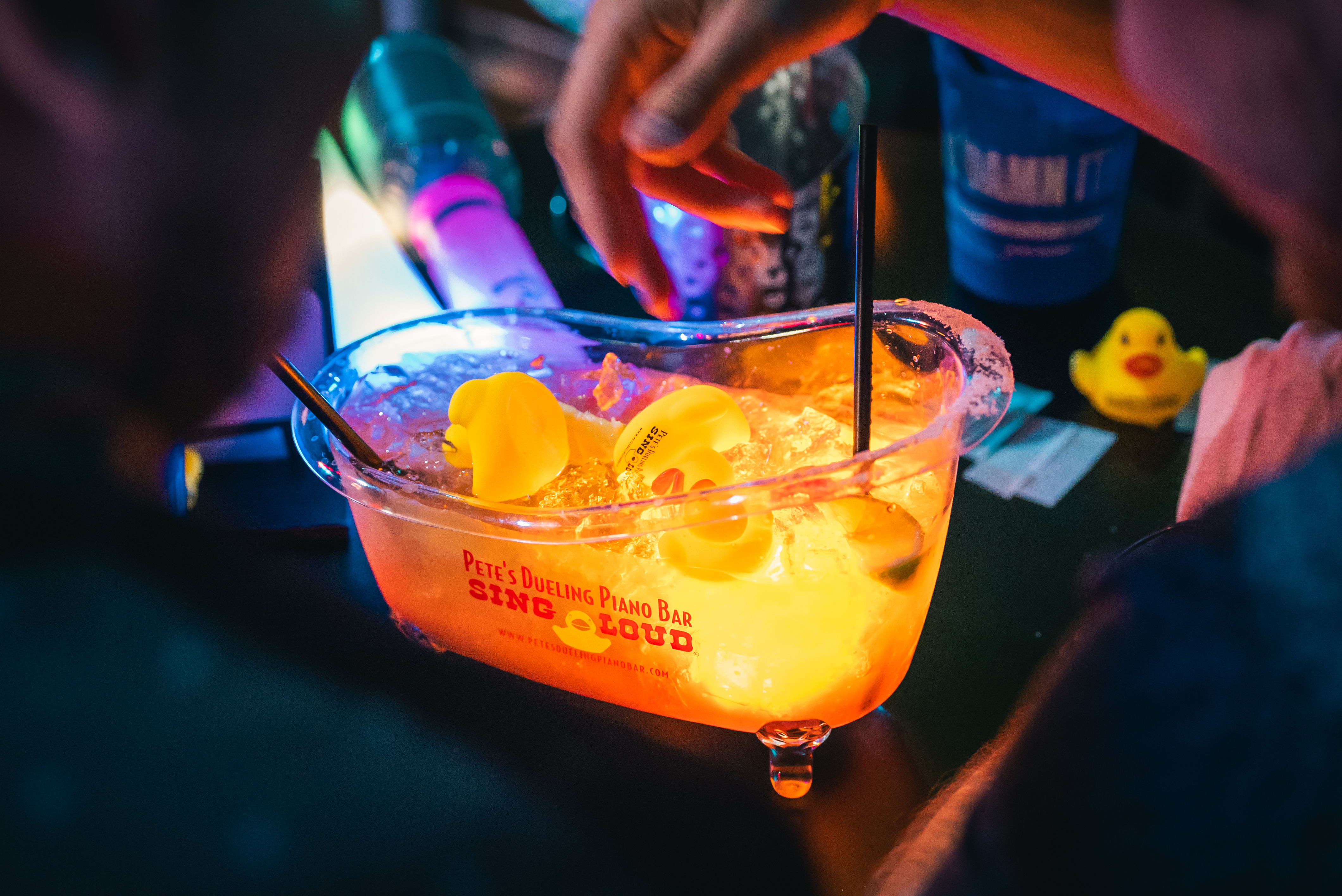 Neon-lit nightlife scene: glowing ice bucket centerpiece on a bar table filled with ice and rubber duckies, colorful straws and a hand reaching in.