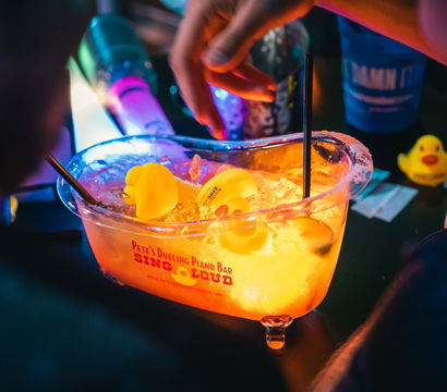 Neon-lit nightlife scene: glowing ice bucket centerpiece on a bar table filled with ice and rubber duckies, colorful straws and a hand reaching in.
