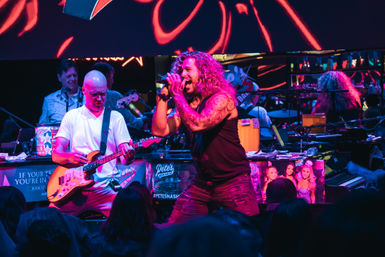 Energetic live band performing under neon pink and blue lights — tattooed curly-haired lead singer belting into a microphone, guitarist playing electric guitar, band and crowd silhouettes in the background.