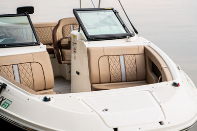 Cozy bow seating of a white motorboat with tan quilted upholstery, open center windshield and white fiberglass deck floating on calm water