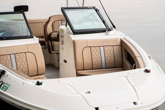 Cozy bow seating of a white motorboat with tan quilted upholstery, open center windshield and white fiberglass deck floating on calm water