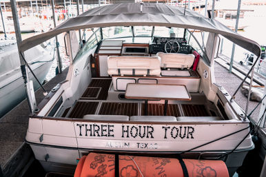 White tour boat docked in a marina slip, aft view with covered cockpit, cushioned bench seating, folding table, helm with steering wheel, and orange life mats.