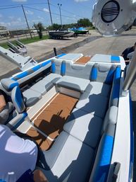 Modern wakeboat interior in a sunny marina: gray and blue cushioned L-shaped seating, brown faux-teak deck panels, wakeboard tower with speakers, and other boats on trailers visible in the background.