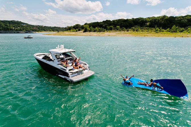Friends enjoying summer boating and lounging on a large blue inflatable mat beside a black-and-white powerboat on a turquoise lake with a tree-lined shore under a sunny sky