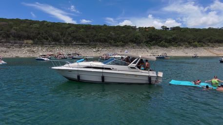 White cabin motorboat anchored on a blue-green lake by a rocky, tree-lined shoreline with people relaxing on a bright blue floating mat and other party boats in the distance under a sunny sky.