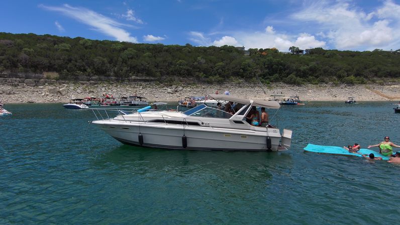 White cabin motorboat anchored on a blue-green lake by a rocky, tree-lined shoreline with people relaxing on a bright blue floating mat and other party boats in the distance under a sunny sky.