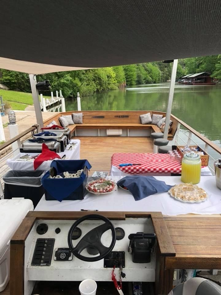 View from the helm of a party-ready pontoon boat on a calm wooded lake, showing wooden deck seating, buffet tables with a red-check tablecloth, trays of appetizers and a lemonade dispenser, with docks and a boathouse along the tree-lined shoreline.