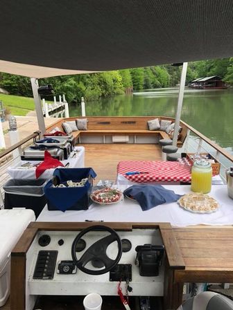 View from the helm of a party-ready pontoon boat on a calm wooded lake, showing wooden deck seating, buffet tables with a red-check tablecloth, trays of appetizers and a lemonade dispenser, with docks and a boathouse along the tree-lined shoreline.