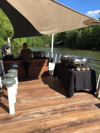 Sunlit riverboat deck with a shade sail, buffet table and drink station, bar stools along the railing and a tree-lined riverbank in the background