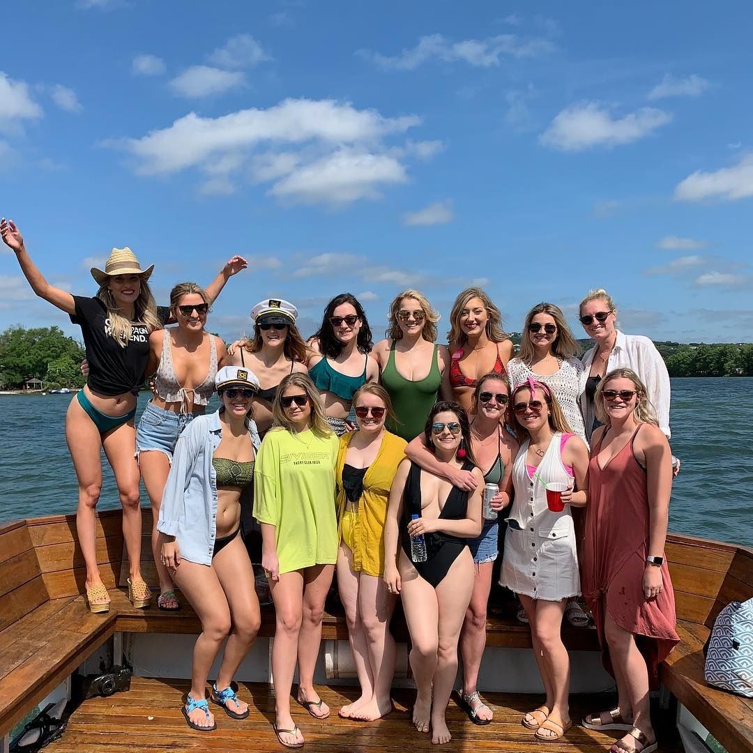 Cheerful group of women on a wooden boat enjoying a sunny lake day in swimsuits and summer outfits under a bright blue sky with scattered clouds