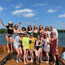 Cheerful group of women on a wooden boat enjoying a sunny lake day in swimsuits and summer outfits under a bright blue sky with scattered clouds