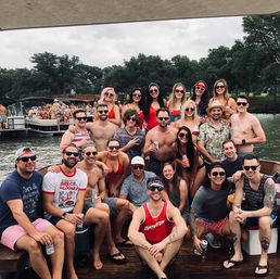 Large group of friends in swimsuits and sunglasses posing on a dock and pontoon boats at a lakeside party, many holding drinks with trees and a cloudy sky in the background.