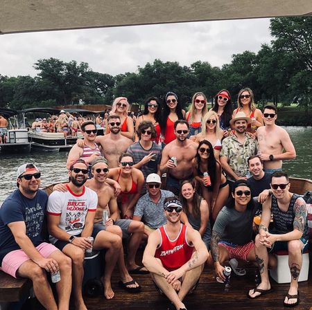 Large group of friends in swimsuits and sunglasses posing on a dock and pontoon boats at a lakeside party, many holding drinks with trees and a cloudy sky in the background.