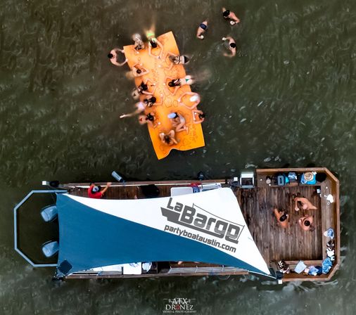Aerial drone view of a lively lake party with a blue-canopied boat, people on a wooden deck and clustered on a bright orange floating mat while others swim nearby.