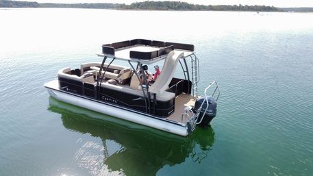 Pontoon boat with cushioned seating and a deck slide floating on a calm green lake, a person relaxing onboard and a tree-lined shoreline in the distance