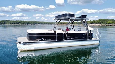 Sleek black-and-white pontoon boat floating on a glassy lake under a sunny blue sky with puffy clouds and a tree-lined shoreline